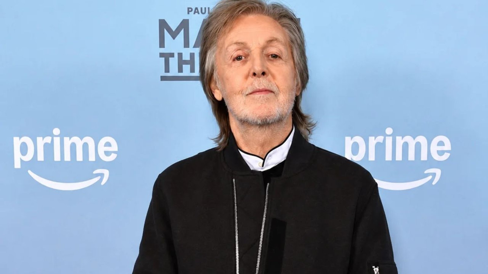 Paul McCartney de jaqueta preta, fundo azul com logotipos Prime Video e PAUL M TH