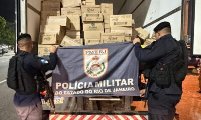 Dois policiais militares seguram bandeira PMERJ, com caixas "brf", "FLORA" em caminhão
