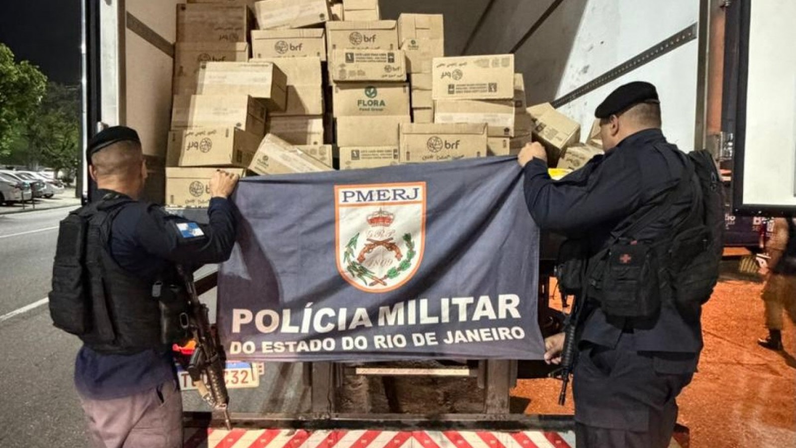 Dois policiais militares seguram bandeira PMERJ, com caixas "brf", "FLORA" em caminhão