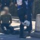 Homem sentado no chão, com mãos para trás, cercado por policiais fardados; um segura capacete branco