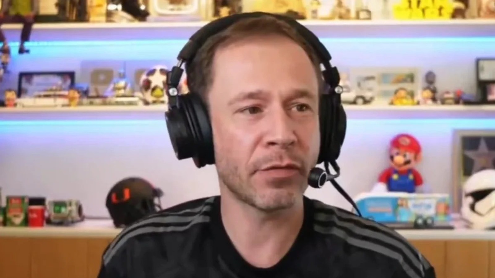 Tiago Leifert usando fones de ouvido e microfone, com fundo de prateleiras iluminadas