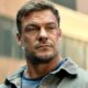 Rosto do ator Alan Ritchson em close-up, com arranhões e barba, vestindo jaqueta