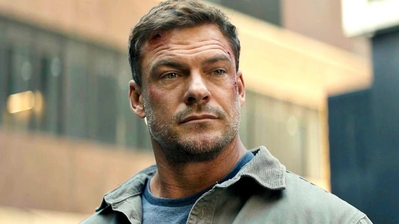 Rosto do ator Alan Ritchson em close-up, com arranhões e barba, vestindo jaqueta