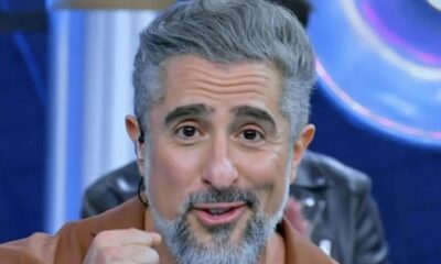 Marcos Mion, cabelo e barba grisalhos, sorrindo, olhando para frente, com earbud em estúdio