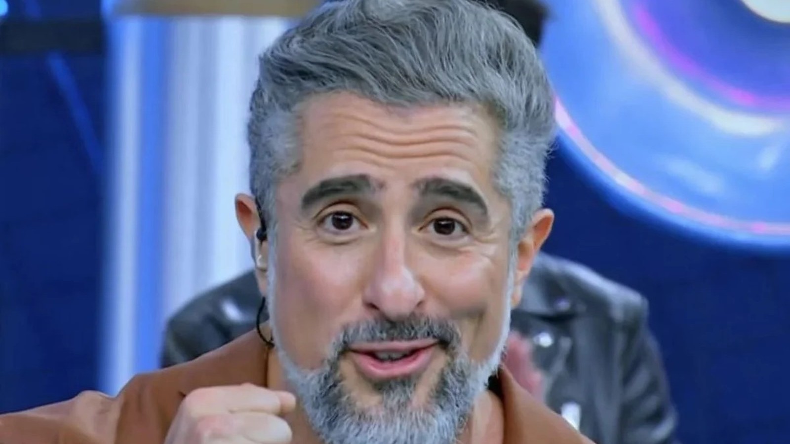 Marcos Mion, cabelo e barba grisalhos, sorrindo, olhando para frente, com earbud em estúdio