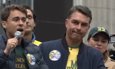 Nikolas Ferreira discursa ao microfone, Flávio Bolsonaro ao lado, mulher com boné político
