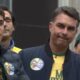 Nikolas Ferreira discursa ao microfone, Flávio Bolsonaro ao lado, mulher com boné político