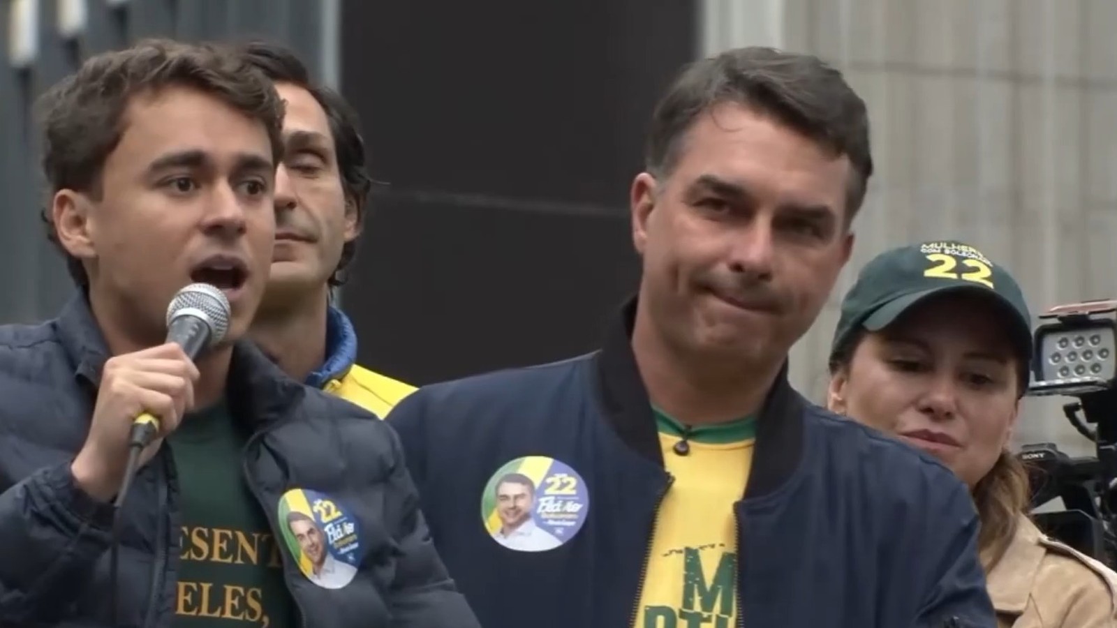 Nikolas Ferreira discursa ao microfone, Flávio Bolsonaro ao lado, mulher com boné político