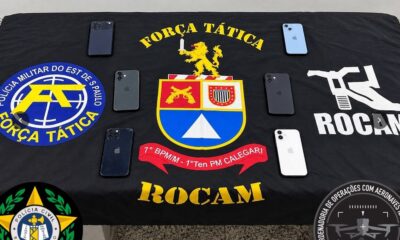 Seis iPhones sobre bandeira preta com emblemas da Polícia Militar e Civil de São Paulo