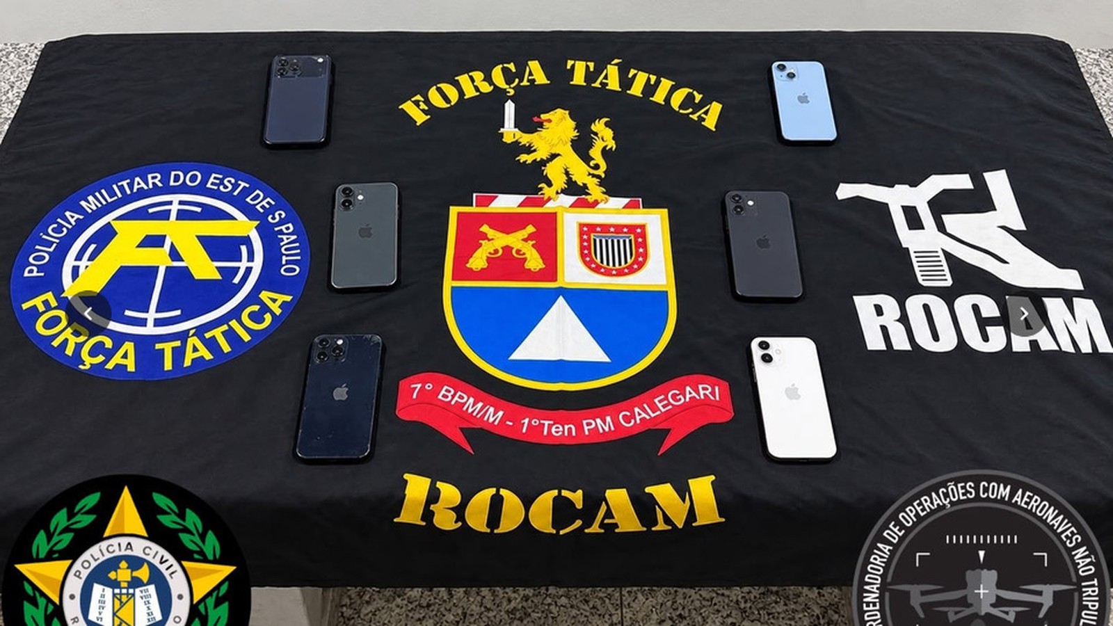 Seis iPhones sobre bandeira preta com emblemas da Polícia Militar e Civil de São Paulo