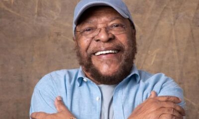 Homem idoso, negro, barbudo, de óculos e boné azul claro sorri com os braços cruzados