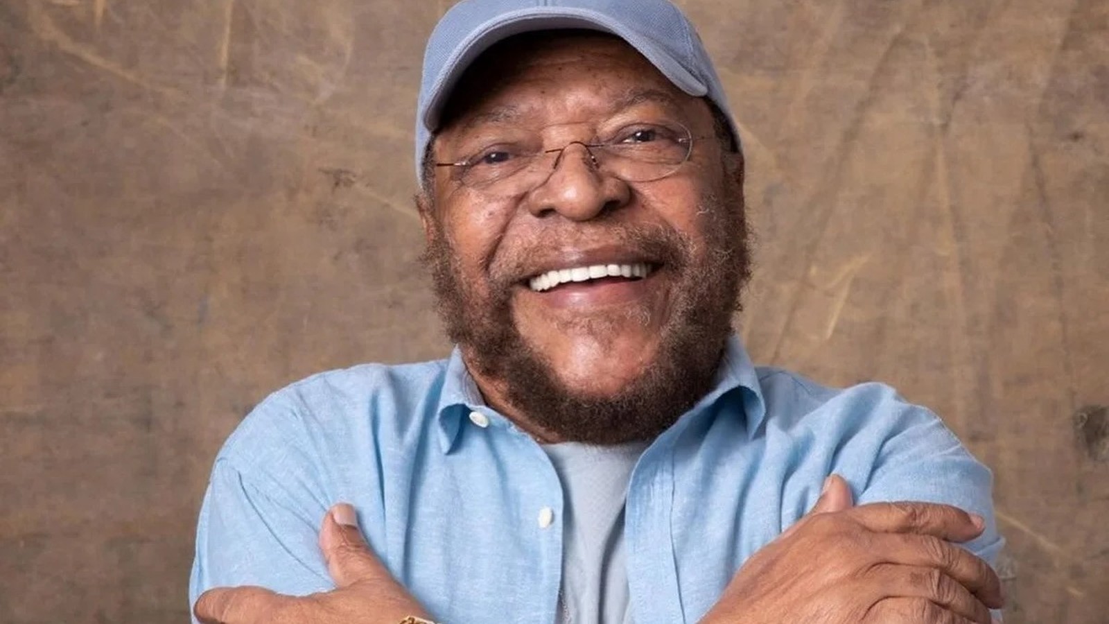 Homem idoso, negro, barbudo, de óculos e boné azul claro sorri com os braços cruzados