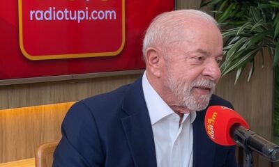 Luiz Inácio Lula da Silva sorrindo, em estúdio de rádio com microfone e tela 'radiotupi.com'