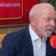Luiz Inácio Lula da Silva sorrindo, em estúdio de rádio com microfone e tela 'radiotupi.com'