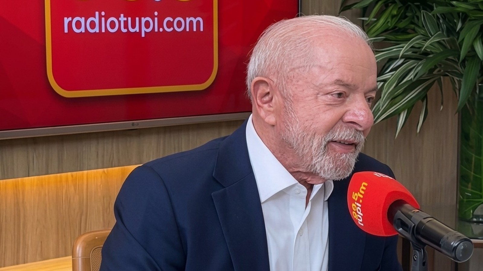 Luiz Inácio Lula da Silva sorrindo, em estúdio de rádio com microfone e tela 'radiotupi.com'
