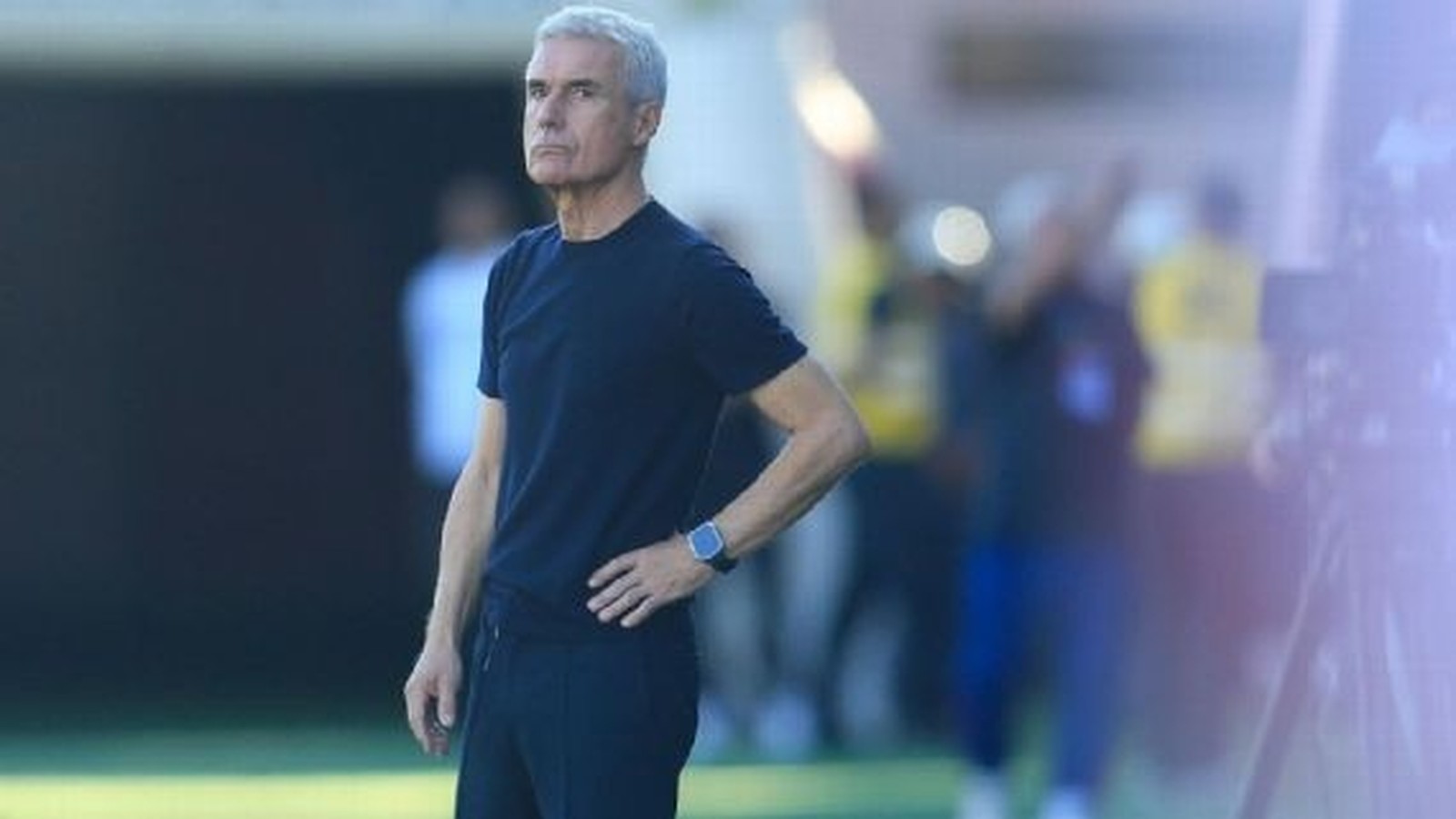 Homem de cabelo grisalho e camiseta azul escura, com relógio no pulso, em campo de futebol