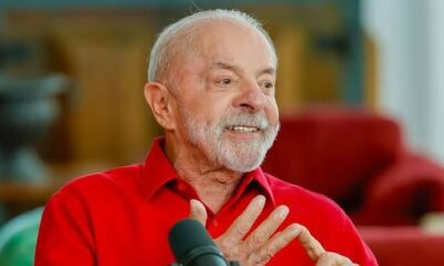 Luiz Inácio Lula da Silva, camisa vermelha, sorrindo e gesticulando em frente a um microfone.