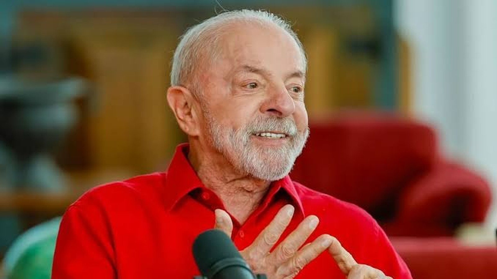 Luiz Inácio Lula da Silva, camisa vermelha, sorrindo e gesticulando em frente a um microfone.
