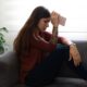 Jovem mulher triste sentada no sofá, mão na testa com celular, planta ao lado