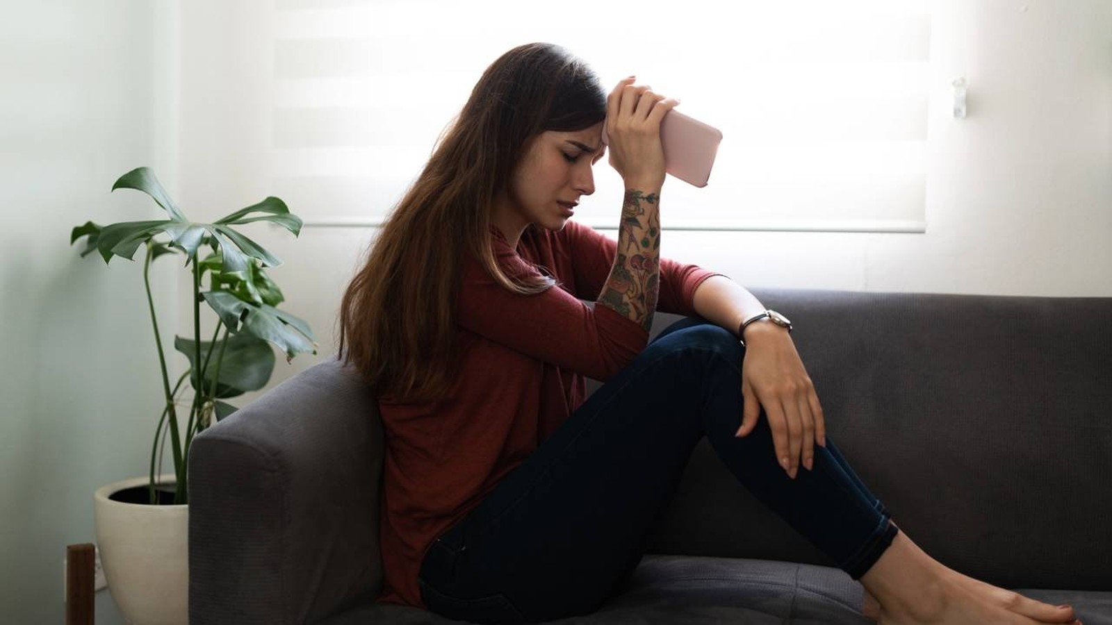 Jovem mulher triste sentada no sofá, mão na testa com celular, planta ao lado