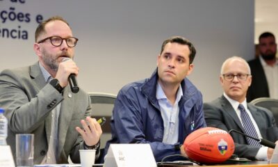 Três homens em reunião, um falando ao microfone, com bola de futebol americano Wilson The Duke Rio