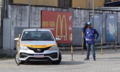 Carro de autoescola prateado em pista de exames, homem em colete azul e cones amarelos