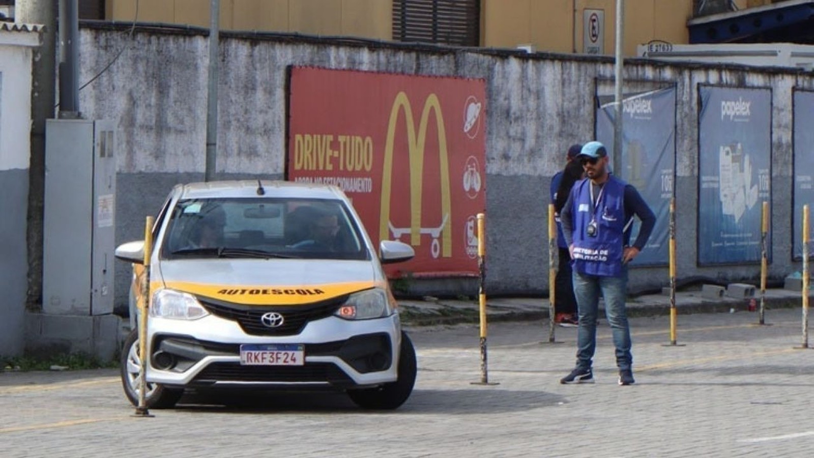 Carro de autoescola prateado em pista de exames, homem em colete azul e cones amarelos