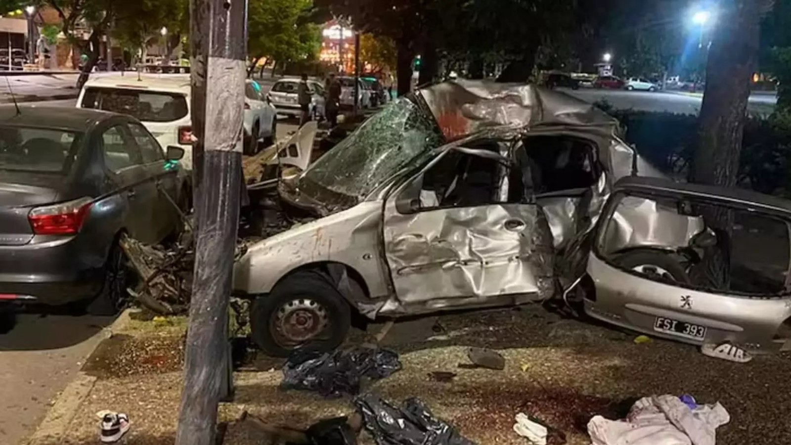Carro prata destruído após colisão, outro veículo danificado e destroços na rua noturna