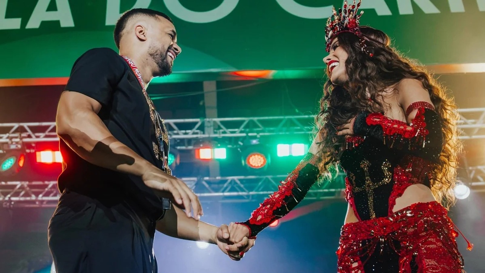 Anitta e Mestre Fafá sorrindo, de mãos dadas, em palco iluminado com luzes coloridas