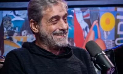 Homem de barba grisalha e camiseta preta sorri, microfone à direita, fundo artístico