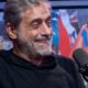 Homem de barba grisalha e camiseta preta sorri, microfone à direita, fundo artístico