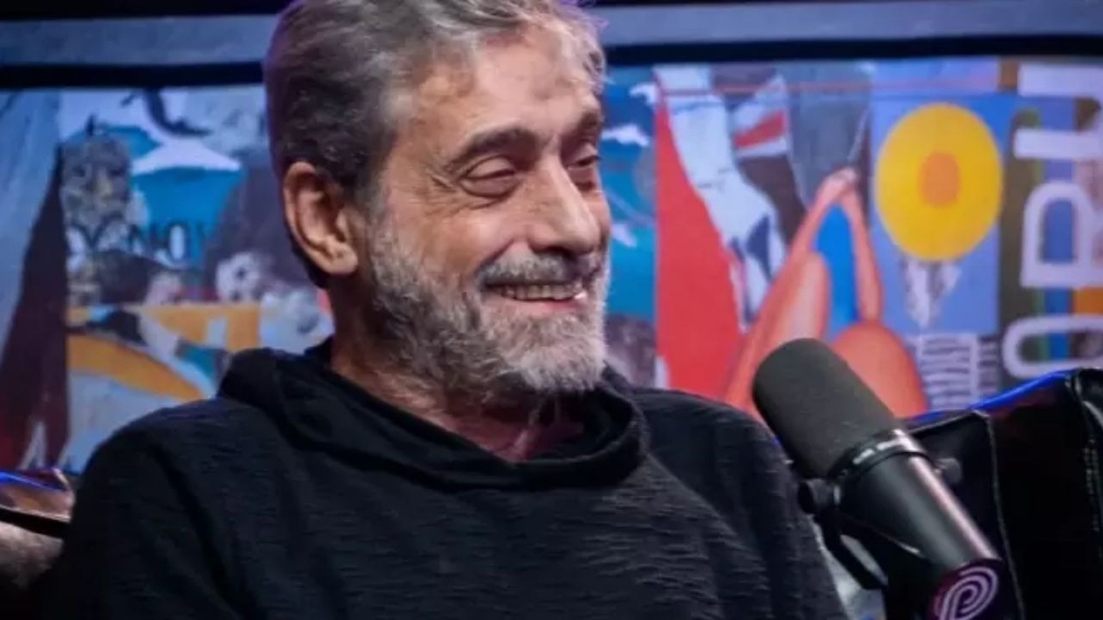 Homem de barba grisalha e camiseta preta sorri, microfone à direita, fundo artístico