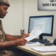 Oficial militar de uniforme lendo papéis em uma mesa com computador e placa 'ADI 7903'