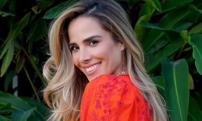 Close-up de mulher sorrindo, cabelo loiro, blusa laranja estampada, fundo de folhas verdes