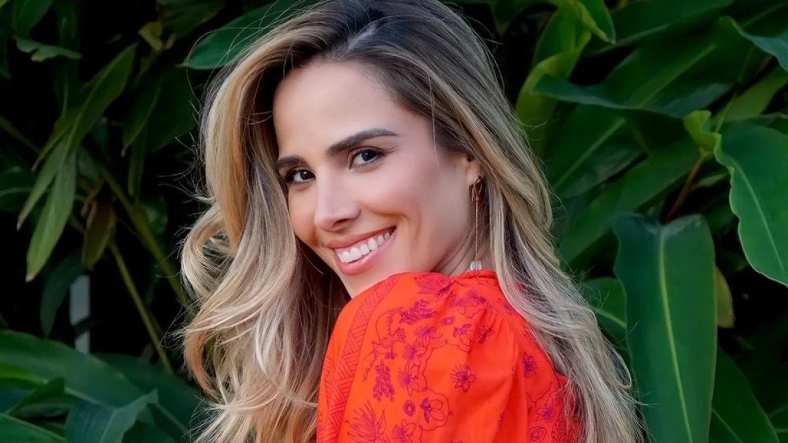 Close-up de mulher sorrindo, cabelo loiro, blusa laranja estampada, fundo de folhas verdes