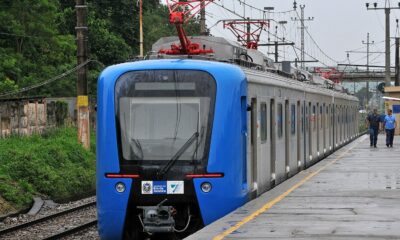 Trem azul e prateado na plataforma de estação com dois homens e vegetação ao lado.