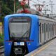 Trem azul e prateado na plataforma de estação com dois homens e vegetação ao lado.