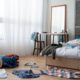 Quarto bagunçado: roupas espalhadas no chão, cama, cadeira; espelho, cortinas brancas