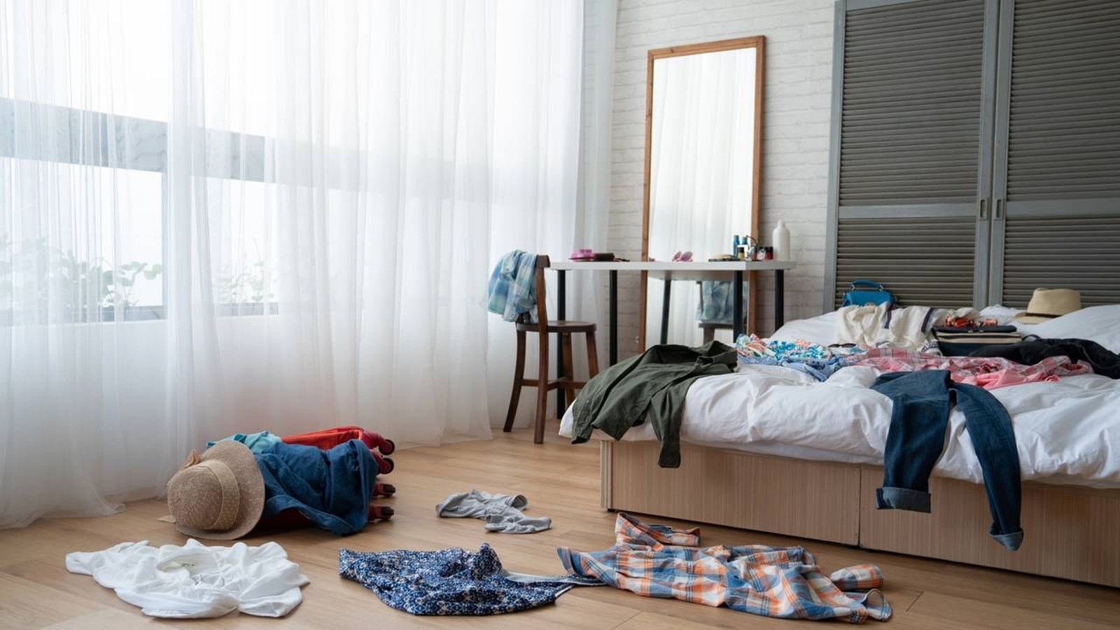 Quarto bagunçado: roupas espalhadas no chão, cama, cadeira; espelho, cortinas brancas