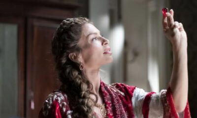 Mulher de perfil com trança, vestido vermelho bordado, segura objeto vermelho translúcido, olhando para cima