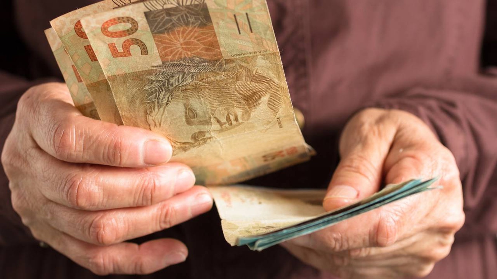 Mãos enrugadas segurando cédulas de 50 reais