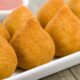 Coxinhas douradas em travessa branca, com molho vermelho e erva verde em fundo desfocado