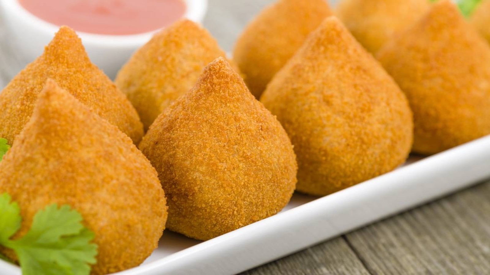 Coxinhas douradas em travessa branca, com molho vermelho e erva verde em fundo desfocado