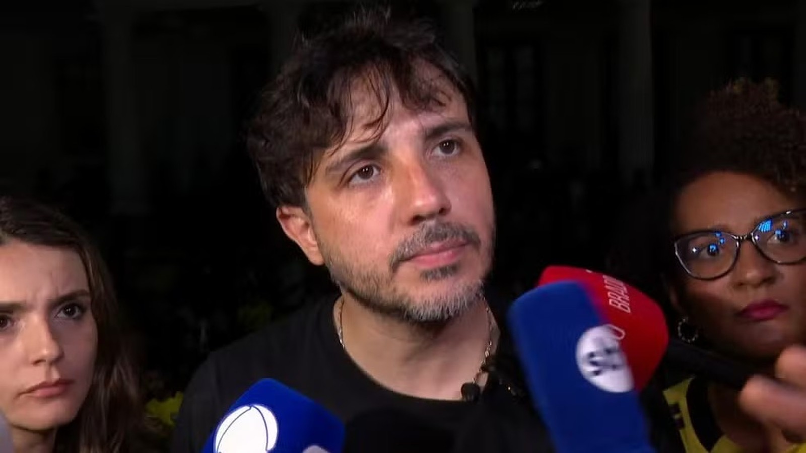 Homem com barba, cabelo escuro, fala para microfones de TV; um deles exibe 'BRAD'