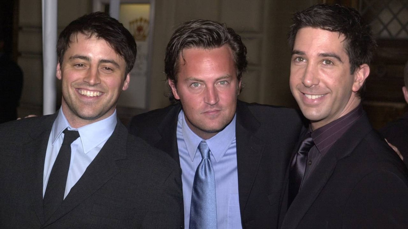 Matt LeBlanc, Matthew Perry e David Schwimmer, sorrindo, em ternos escuros, posando.