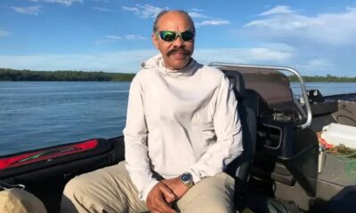 Homem de óculos escuros, bigode, camisa clara, sentado em barco. Rio e floresta ao fundo