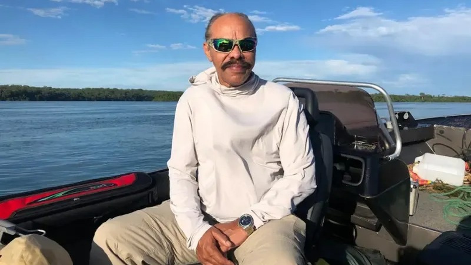Homem de óculos escuros, bigode, camisa clara, sentado em barco. Rio e floresta ao fundo