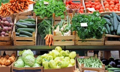Caixas de madeira cheias de vegetais frescos como cenouras, beterrabas, abobrinhas, tomates e pimentões com etiquetas de preço "CIKLA" e "5"
