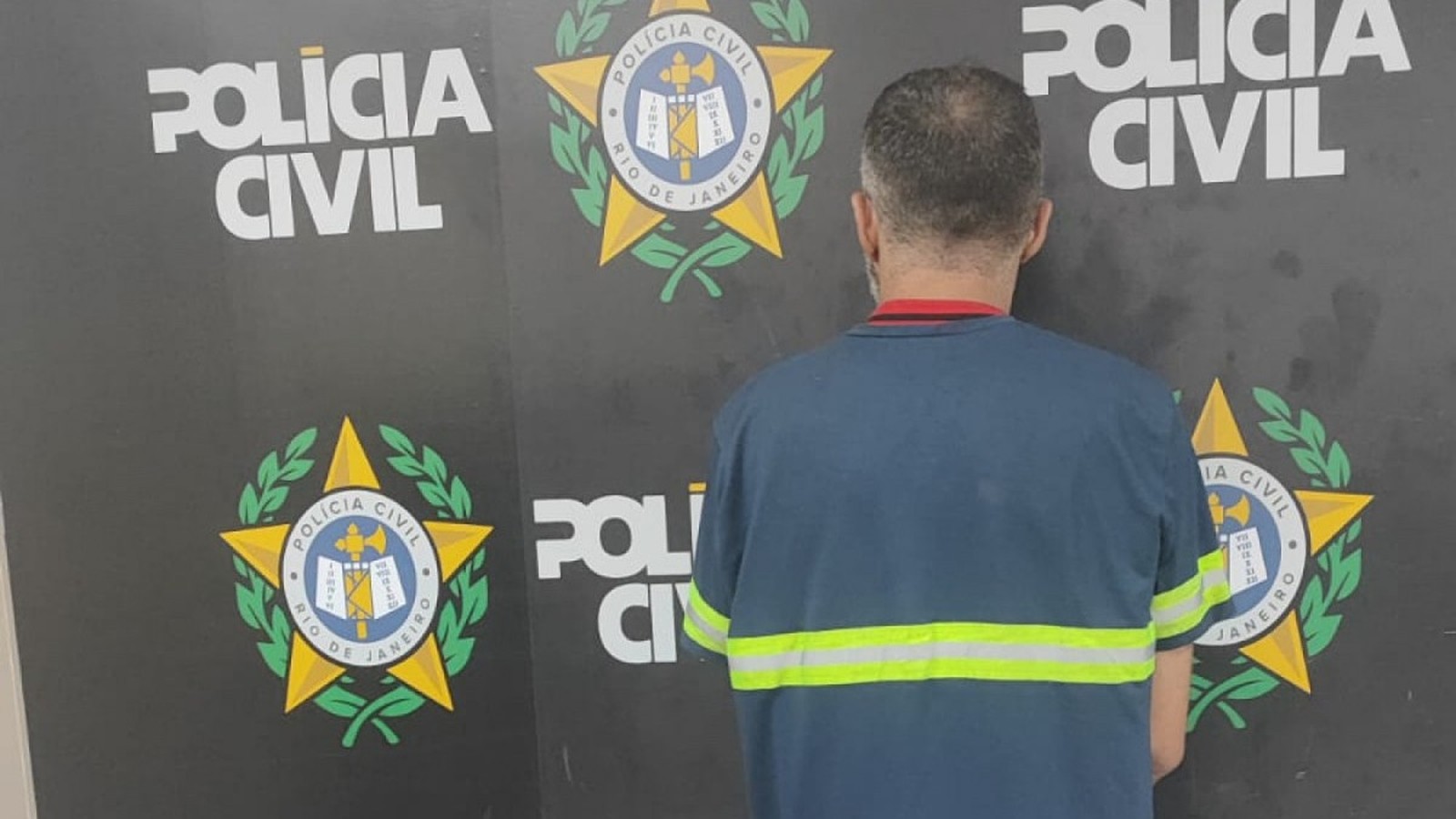 Homem de costas em camiseta azul com faixas refletivas, em frente ao logo da Polícia Civil do Rio de Janeiro