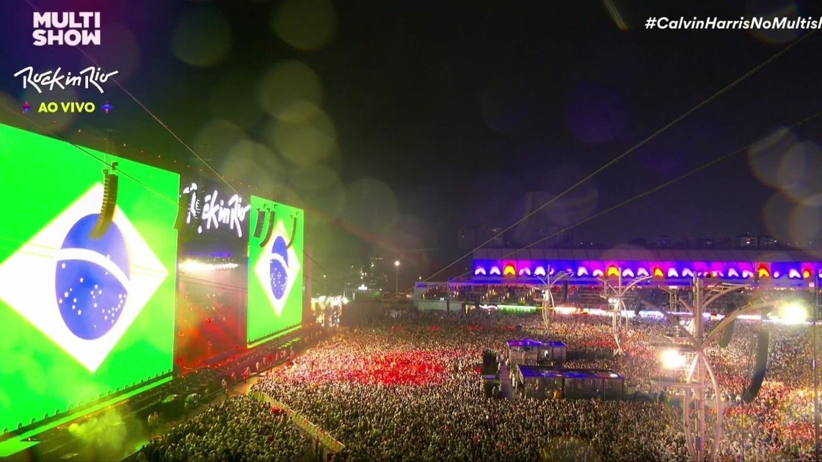Vista aérea noturna do palco principal do Rock in Rio com telões exibindo a bandeira do Brasil e texto
